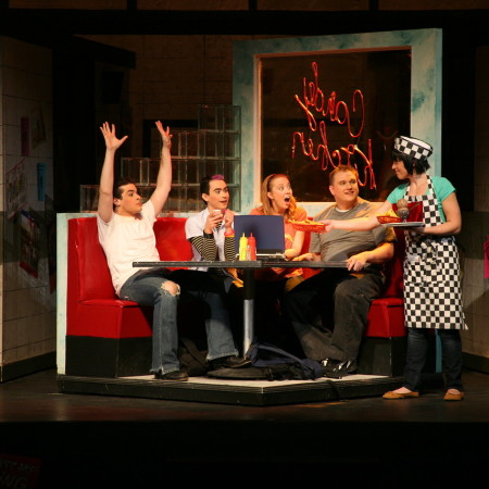 Four actors sit in a diner booth set on stage while a waitress in a checkered apron serves them.
