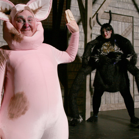 Wilbur waves his hand as Charlotte watches from the background near a barn on the set of Charlottes web.