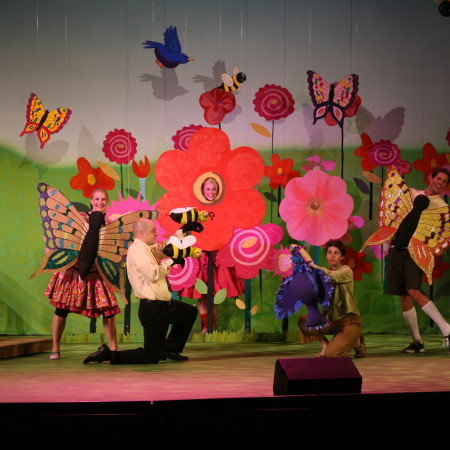 Four actors hold butterfly, bumblebee and bird puppets in front of a bright oversized flower garden set piece on stage.