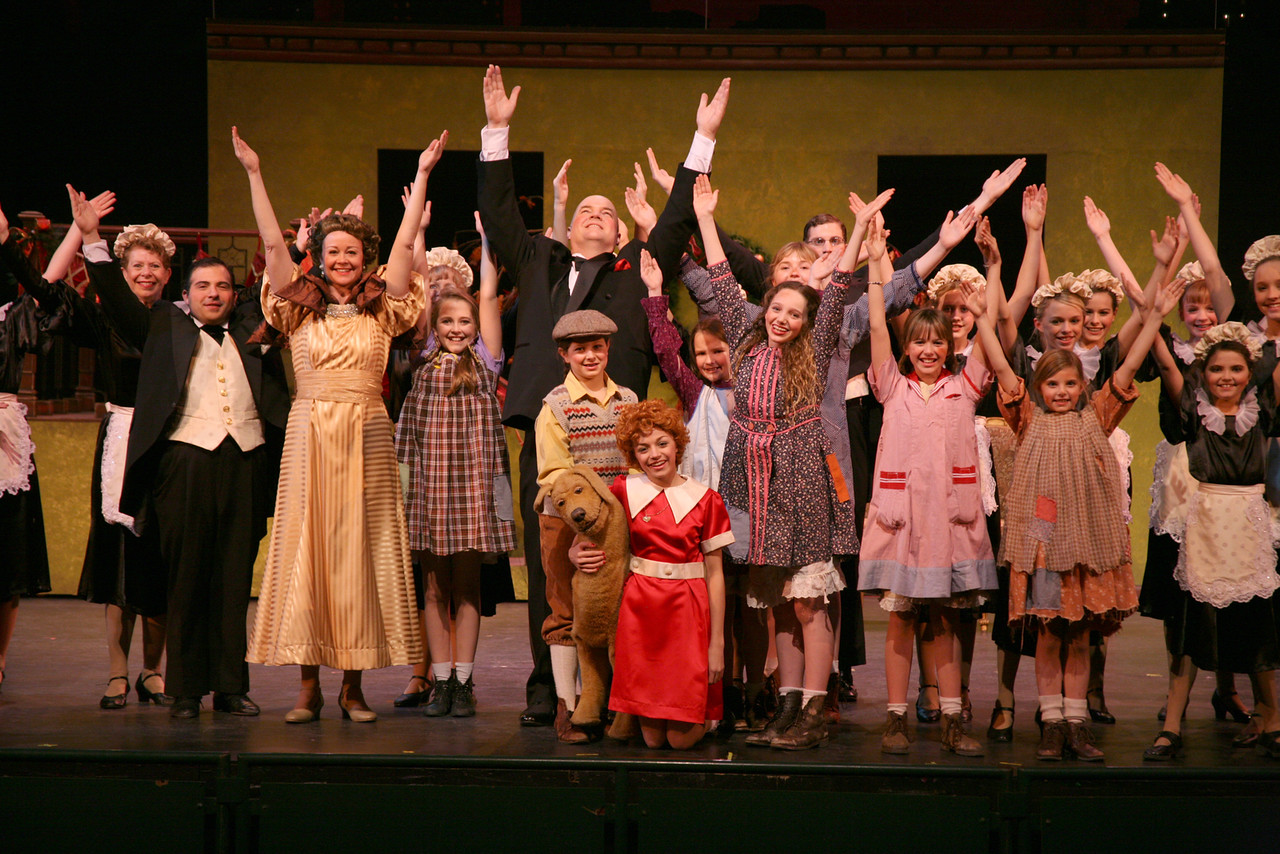 The cast of Annie all hold their arms overhead while surrounding Annie and her dog on stage.