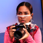 Nivi Varanasi as Vita in HOW TO BUILD AN ARK. PHOTO DESCRIPTION: A youth actor wearing a pink shirt holds a camera, about to take a photo.