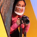 Nivi Varanasi as Vita in HOW TO BUILD AN ARK. PHOTO DESCRIPTION: A youth actor wearing a pink shirt stands behind the trunk of a tree, holding a camera.