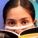 Nivi Varanasi as Vita in HOW TO BUILD AN ARK. PHOTO DESCRIPTION: A youth actor wearing a clear face mask peeks over the pages of a National Geographic magazine.