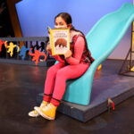 Nivi Varanasi as Vita in HOW TO BUILD AN ARK. PHOTO DESCRIPTION: A youth actor wearing a pink shirt, plaid vest, pink leggings, and yellow high-top tennis shoes sits on a turquoise playground slide while reading a National Geographic magazine. The cover image shows an armadillo with a headline about the Photo Ark.