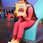 Nivi Varanasi as Vita in HOW TO BUILD AN ARK. PHOTO DESCRIPTION: A youth actor wearing a pink shirt, plaid vest, pink leggings, and yellow high-top tennis shoes sits on a turquoise playground slide while reading a National Geographic magazine. The cover image shows an armadillo with a headline about the Photo Ark.