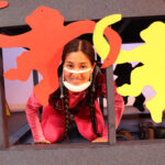 Nivi Varanasi as Vita in HOW TO BUILD AN ARK. PHOTO DESCRIPTION: A smiling youth actor wearing a pink shirt peeks out under the arm of stylized red and yellow monkey cut outs.
