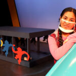 Nivi Varanasi as Vita in HOW TO BUILD AN ARK. PHOTO DESCRIPTION: A youth actor wearing a pink shirt leans alongside a turquoise playground slide. A black platform decorated with red, yellow and blue monkey figures can be seen behind her.