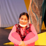 Nivi Varanasi as Vita in HOW TO BUILD AN ARK. PHOTO DESCRIPTION: A youth actor wearing a plaid vest, pink shirt, pink leggings, and yellow high-top tennis shoes sits cross-legged beside a large yellow set piece reminiscent of the National Geographic magazine border.