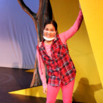 Nivi Varanasi as Vita in HOW TO BUILD AN ARK. PHOTO DESCRIPTION: A youth actor wearing a plaid vest, pink shirt, and pink leggings stands beside a large yellow set piece reminiscent of the National Geographic magazine border.