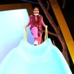Nivi Varanasi as Vita in HOW TO BUILD AN ARK. PHOTO DESCRIPTION: A youth actor wearing a plaid vest, pink shirt, and pink leggings stands at the top of a playground slide.