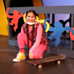Nivi Varanasi as Vita in HOW TO BUILD AN ARK. PHOTO DESCRIPTION: A youth actor wearing a plaid vest, pink shirt and pink kneels by a skateboard. In the background can be seen a platform decorated with brightly-colored cutouts of monkey figures.