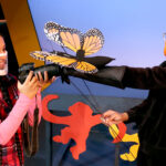 Nivi Varanasi as Vita and Tamia Renée as the Monarch Butterfly in HOW TO BUILD AN ARK. PHOTO DESCRIPTION: A youth actor wearing a plaid vest, pink shirt and pink leggings looks at an orange and black Monarch butterfly puppet that has landed on the lens of her camera. The butterfly puppeteer wears all black clothing and an orange mask. In the background can be seen a tree and a platform decorated with brightly-colored cutouts of monkey figures.