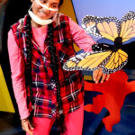 Nivi Varanasi as Vita in HOW TO BUILD AN ARK. PHOTO DESCRIPTION: A youth actor wearing a plaid vest, pink shirt and pink leggings looks at an orange and black Monarch butterfly puppet. In the background can be seen a tree and a platform decorated with brightly-colored cutouts of monkey figures.