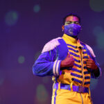 Photo description: A male actor is wearing a blue military-inspired jacket over a yellow and blue jumpsuit with bright blue boots and a blue mask. His hands are holding his lapels as he stands in front of a background of blue bokeh light bubbles.