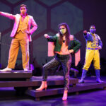 Photo description: Three actors strike a dramatic pose with their arms outstretched and their hands in fists. A male actor wearing a sparkly pink blazer over a gold vest and pants with a pink button-up shirt has his arm stretched to the left. Another male actor is wearing a blue military-inspired jacket over a yellow and blue jumpsuit with bright blue boots and a blue mask; his arm is stretched out to the camera. A female actor is wearing a greet jacket embellished with pink sequins and long pink fringe along the sleeves and back of the jacket. She wears striped grey pants with pink sequin trim and pink high-heeled boots.
