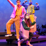 Photo description: Three actors strike a dramatic pose. A male actor wearing a sparkly pink blazer over a gold vest and pants with a pink button-up shirt has his arm stretched upwards with his hand in a fist. Another male actor is wearing a blue military-inspired jacket over a yellow and blue jumpsuit with bright blue boots and a blue mask. He is flexing both of his arms in a power pose. A female actor is wearing a greet jacket embellished with pink sequins and long pink fringe along the sleeves and back of the jacket. She wears striped grey pants with pink sequin trim and pink high-heeled boots as she poses sideways with both hands in fists.