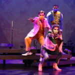 Photo description: Three actors strike a dramatic pose with their arms outstretched. A male actor wearing a sparkly pink blazer over a gold vest and pants with a pink button-up shirt has his arm stretched toward the camera. Another male actor is wearing a blue military-inspired jacket over a yellow and blue jumpsuit with bright blue boots and a blue mask. A female actor is wearing a greet jacket embellished with pink sequins and long pink fringe along the sleeves and back of the jacket. She wears striped grey pants with pink sequin trim and pink high-heeled boots as she reaches out with both hands toward the camera