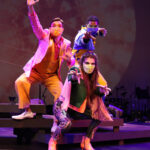 Photo description: Three actors strike a dramatic pose with their arms outstretched. A male actor wearing a sparkly pink blazer over a gold vest and pants with a pink button-up shirt has his arm stretched upwards. Another male actor is wearing a blue military-inspired jacket over a yellow and blue jumpsuit with bright blue boots and a blue mask; his arm is stretched out to the camera. A female actor is wearing a greet jacket embellished with pink sequins and long pink fringe along the sleeves and back of the jacket. She wears striped grey pants with pink sequin trim and pink high-heeled boots as she reaches out with both hands toward the camera