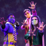 Photo description: Three actors strike a dramatic pose with their arms outstretched. A male actor is wearing a blue military-inspired jacket over a yellow and blue jumpsuit with bright blue boots and a blue mask. A female actor is wearing a greet jacket embellished with pink sequins and long pink fringe along the sleeves and back of the jacket. She wears striped grey pants with pink sequin trim and pink high-heeled boots. Another male actor wears a sparkly pink blazer over a gold vest and pants with a pink button-up shirt.