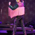 Photo description: A female actor is wearing a greet jacket embellished with pink sequins and long pink fringe along the sleeves and back of the jacket. She wears striped grey pants with pink sequin trim and pink high-heeled boots. Her back is to the camera and her arms are flexed in a position of strength. She looks over her shoulder to the camera in a powerful pose.