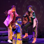 Photo description: Three actors strike a dramatic pose with their arms outstretched. A male actor wears a sparkly pink blazer over a gold vest and pants with a pink button-up shirt. Another male actor is wearing a blue military-inspired jacket over a yellow and blue jumpsuit with bright blue boots and a blue mask. A female actor is wearing a greet jacket embellished with pink sequins and long pink fringe along the sleeves and back of the jacket. She wears striped grey pants with pink sequin trim and pink high-heeled boots.