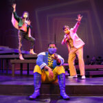 Photo description: Three actors strike a dramatic pose on a set of stairs with their arms outstretched. A female actor is wearing a greet jacket embellished with pink sequins and long pink fringe along the sleeves and back of the jacket. She wears striped grey pants with pink sequin trim and pink high-heeled boots. Another male actor wears a sparkly pink blazer over a gold vest and pants with a pink button-up shirt. A second male actor is sitting on the step closest to the camera. He is wearing a blue military-inspired jacket over a yellow and blue jumpsuit with bright blue boots and a blue mask.