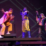 Photo description: Three actors strike a dramatic pose with their arms outstretched. A male actor wears a sparkly pink blazer over a gold vest and pants with a pink button-up shirt. Another male actor is wearing a blue military-inspired jacket over a yellow and blue jumpsuit with bright blue boots and a blue mask. A female actor is wearing a greet jacket embellished with pink sequins and long pink fringe along the sleeves and back of the jacket. She wears striped grey pants with pink sequin trim and pink high-heeled boots.