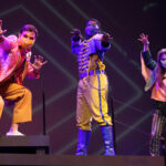 Photo description: Three actors strike a dramatic pose with their arms outstretched. A male actor wears a sparkly pink blazer over a gold vest and pants with a pink button-up shirt. Another male actor is wearing a blue military-inspired jacket over a yellow and blue jumpsuit with bright blue boots and a blue mask. A female actor is wearing a greet jacket embellished with pink sequins and long pink fringe along the sleeves and back of the jacket. She wears striped grey pants with pink sequin trim and pink high-heeled boots.